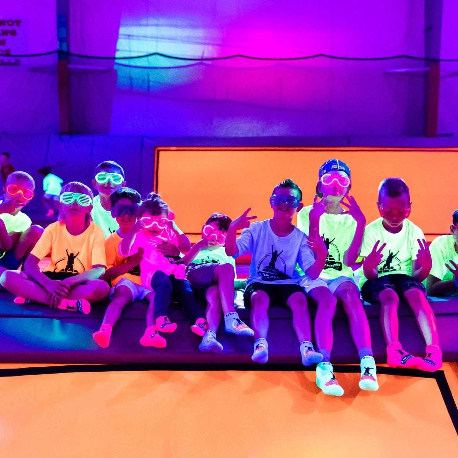 A group of kids striking playful poses in glow glasses under purple and orange trampoline park lights